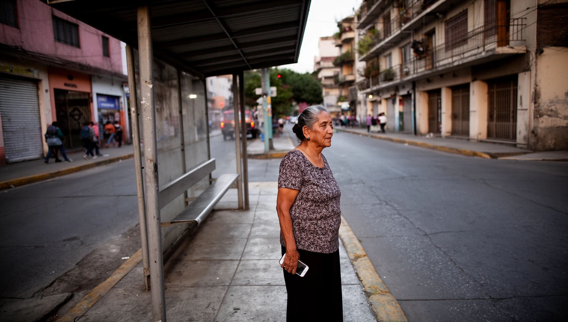 An older woman stands calmly at a bus stop, looking at her smartphone with a confident expression.