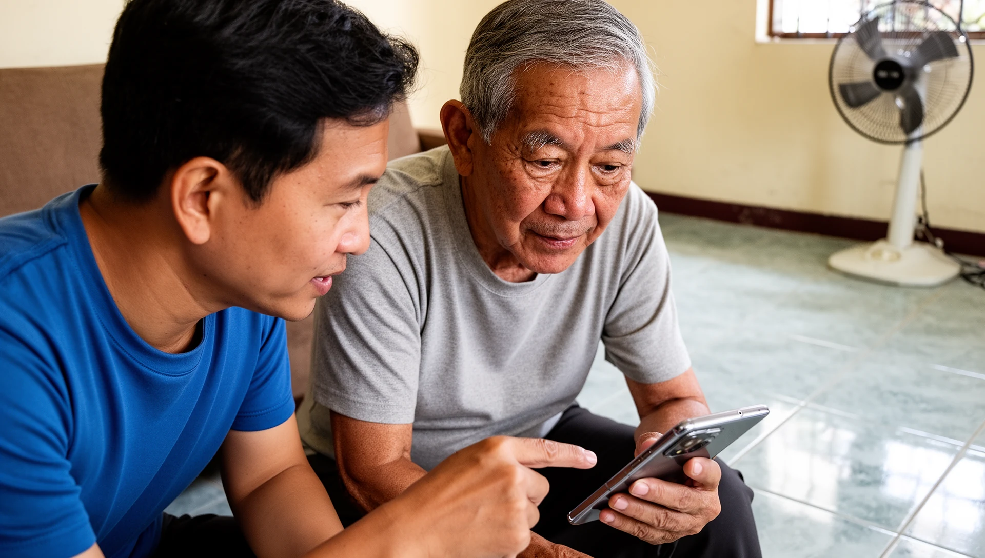 An adult child gently points to a smartphone held by an older Filipino man, who looks at the screen with an expression of understanding and trust, in a home living room.